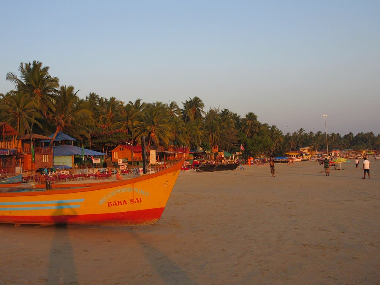 A fishing boat drawn up on the sand at sunset, palms and beach shacks behind, Goa